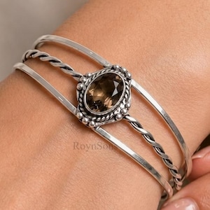 Puede incluir: Un brazalete de plata con una piedra preciosa de cuarzo ahumado de corte ovalado central engastada en un marco decorativo de cuentas. El brazalete consta de tres bandas, incluyendo dos hilos de plata lisos y uno trenzado, con el texto RoynSon.