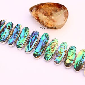 Puede incluir: Pulsera de plata con piedras ovaladas de concha de abulón iridiscente en tonos azules, verdes y morados. Una piedra pulida se encuentra encima de la pulsera. El texto "Roynsons" es visible. Una joya llamativa.