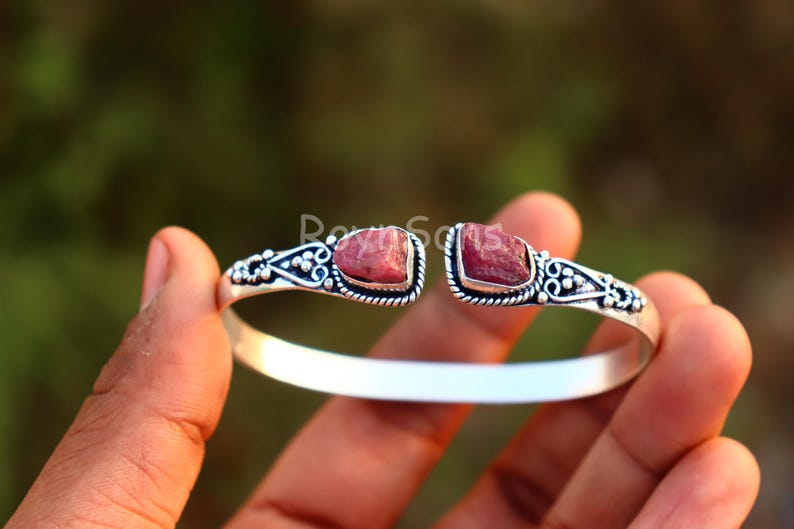 K&ouml;nnte beinhalten: Silberfarbenes Armreif-Armband mit zwei rohen, r&ouml;tlich-rosa Edelsteinen. Das Armband hat verzierte Details um die Edelsteine. Das offene Design erm&ouml;glicht ein einfaches Tragen. Ein einzigartiges Schmuckst&uuml;ck.