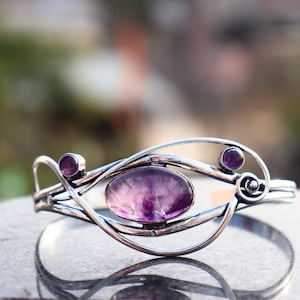 Puede incluir: Brazalete plateado con tres piedras de amatista. La piedra central es ovalada, las otras dos son redondas. El brazalete tiene un diseño calado decorativo con líneas curvas y espirales.