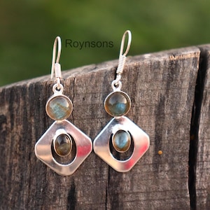 May include: A pair of silver earrings with a geometric design. Each earring features a round, iridescent gemstone at the top and a smaller oval gemstone within a diamond-shaped frame. The earrings are displayed on a weathered wooden surface.
