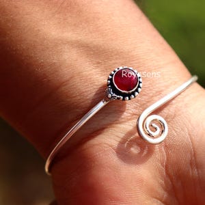 Puede incluir: Brazalete plateado con una piedra preciosa roja en el centro. El brazalete presenta un diseño en espiral en un extremo y un engaste circular alrededor de la piedra preciosa. La piedra preciosa es de color rojo intenso.