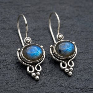 May include: A pair of silver drop earrings featuring oval-shaped labradorite gemstones with iridescent blue flashes. The metal settings include decorative scrollwork and small silver spheres at the base, suspended from curved silver ear wires.
