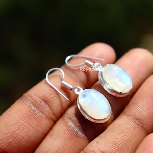 May include: A pair of oval-shaped moonstone earrings with silver hooks. The moonstones have a pearly, iridescent sheen, reflecting light with a soft, ethereal glow. The earrings are displayed against a blurred background.