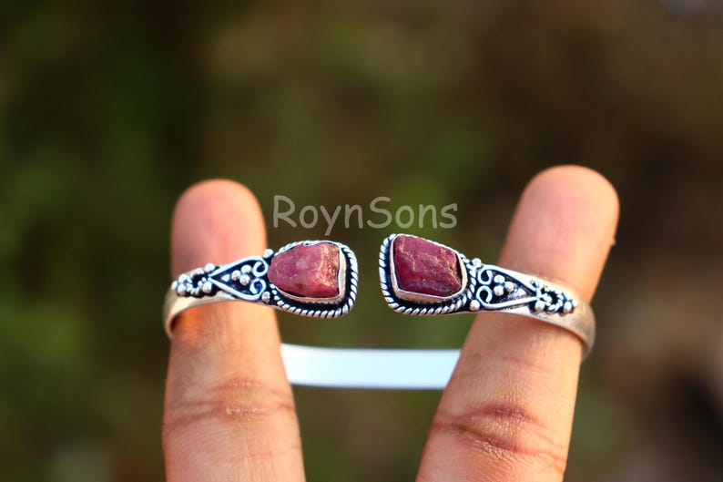 K&ouml;nnte beinhalten: Silberfarbenes Armreif-Armband mit zwei rohen, r&ouml;tlich-rosa Edelsteinen. Das offene Armband hat verzierte Details um die Steine. Die Edelsteine sind in eine dekorative Silberfassung gesetzt und schaffen ein einzigartiges Schmuckst&uuml;ck.