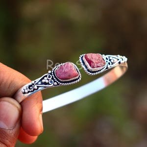 K&ouml;nnte beinhalten: Ein silberfarbenes Armreif-Armband mit zwei rohen, rosa Edelsteinen. Das Armband hat verzierte Details um die Steine und entlang des Bandes. Die Edelsteine sind leuchtend rosa und in eine dekorative silberne Fassung gesetzt.