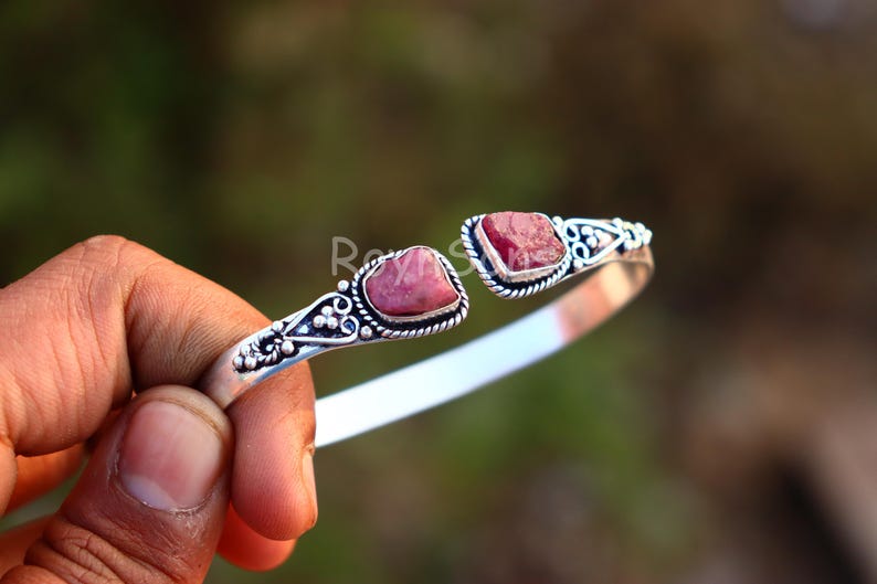 K&ouml;nnte beinhalten: Ein silberfarbenes Armreif-Armband mit zwei rohen, rosa Edelsteinen. Das Armband hat verzierte Details um die Steine und entlang des Bandes. Die Edelsteine sind leuchtend rosa und in eine dekorative silberne Fassung gesetzt.
