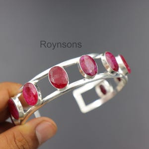 Könnte beinhalten: Ein silbernes Armreif-Armband mit ovalen, tiefroten Edelsteinen. Das Armband hat ein offenes Design mit den Edelsteinen in einzelnen Fassungen. Das Wort "Roynsons" ist im Bild sichtbar.