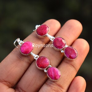 May include: A pair of silver earrings with oval and round, bright pink gemstones. The gemstones are set in a silver metal setting and connected by silver links. The earrings are displayed on a hand.