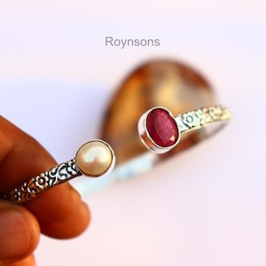 Op de afbeelding: Zilveren manchetarmband met een bloemmotief. De armband heeft een ronde, crèmekleurige parel en een rode robijn edelsteen. Het woord "Roynsons" staat boven de armband.