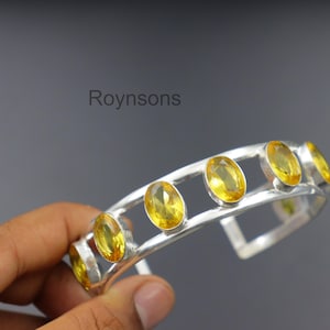 Op de afbeelding: Een zilveren manchetarmband met zeven ovale, gele edelstenen. De armband heeft een modern, open ontwerp met de edelstenen in individuele bezels gezet. Het woord "Roynsons" is zichtbaar op de achtergrond.