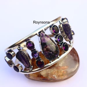 Pode incluir: Uma pulseira de punho prateada com várias pedras preciosas roxas. A pulseira tem um design decorativo vazado e é adornada com pedras ovais, em forma de gota e de formato irregular. A palavra "Roynsons" está impressa na pulseira.