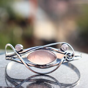 Könnte beinhalten: Ein silberfarbenes Armband mit einem Rosenquarz-Mittelstück und zwei kleineren Rosenquarz-Akzenten. Das Armband hat ein offenes Design mit geschwungenen Linien und eine polierte Oberfläche. Der Rosenquarz hat einen zarten rosa Farbton.