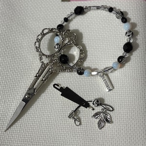 May include: A silver-toned embroidery scissor with ornate handles, a beaded bracelet with black and clear beads, and a small charm with the word "BLESSED". A pink measuring tape shows 3 inches.