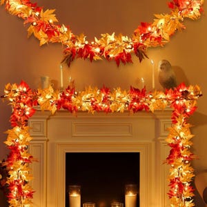 May include: A decorative garland featuring artificial autumn maple leaves in shades of red, orange, and yellow, illuminated by small warm white string lights. The garland is draped across a fireplace mantel and hangs down both sides.