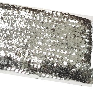 May include: A roll of silver sequin trim featuring a dense arrangement of small, reflective, circular metallic discs attached to a white fabric backing. The trim is wound tightly around a rectangular white cardboard spool for storage and display.