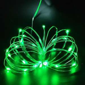 May include: A string of thin, flexible wire lights emitting a bright green glow. The lights are arranged in a loose, circular bundle with multiple individual LED bulbs spaced along the wire, creating a vibrant, luminous display against a dark background.