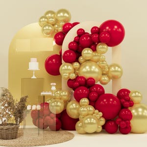 May include: A festive display featuring red and gold balloons of varying sizes, arranged in an arch and clustered formations. A small white cake with pink frosting and other decorative elements are also present, creating a celebratory atmosphere.
