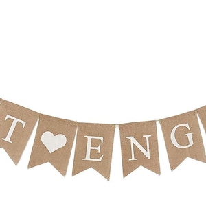 May include: A banner with the words "JUST ENGAGED" in white letters on individual tan pennants. A white heart is between the words. The banner is strung on a thin, silver-colored string.