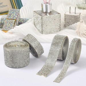 May include: Several rolls of silver rhinestone ribbon featuring a dense grid of small, reflective crystals. The adhesive-backed strips are shown in various widths, with some partially unrolled to display the flexible, sparkling surface texture.