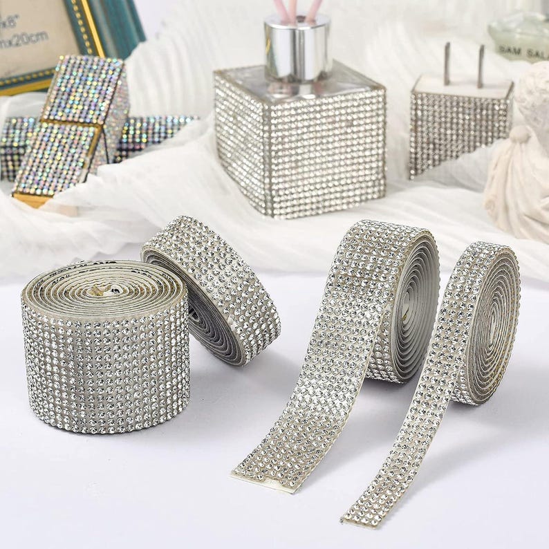 May include: Several rolls of silver rhinestone ribbon featuring a dense grid of small, reflective crystals. The adhesive-backed strips are shown in various widths, with some partially unrolled to display the flexible, sparkling surface texture.