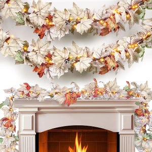 May include: A decorative garland featuring artificial maple leaves in shades of cream, beige, and burnt orange, interspersed with small, warm-glowing LED string lights. The garland is draped across the top of a white fireplace mantel.