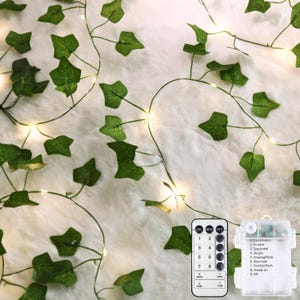 May include: A string of artificial green ivy leaves intertwined with small warm white LED fairy lights. The set includes a rectangular battery pack and a remote control with buttons labeled 1 through 8 for various lighting modes.
