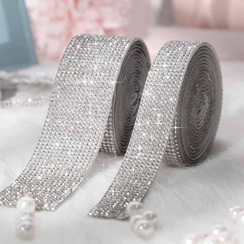 May include: Two rolls of silver rhinestone ribbon featuring a dense grid of small, reflective crystals. The flexible trim has a metallic finish and a sparkling appearance, with each roll coiled tightly to display the continuous crystal texture.