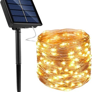 May include: A solar-powered string light set featuring a black plastic ground stake with a rectangular solar panel and a coiled length of thin copper wire adorned with numerous small, glowing warm white LED lights.