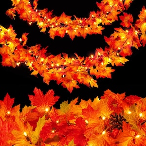 May include: A decorative garland featuring artificial maple leaves in shades of orange, red, and yellow, illuminated by small warm white string lights. The foliage is arranged in a dense, layered pattern against a solid black background.