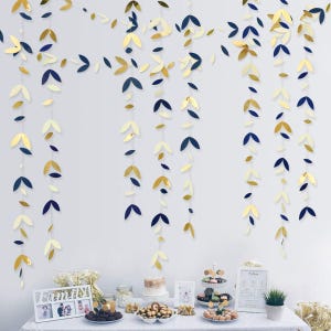 May include: Decorative leaf garlands in gold, navy blue, and cream colors hang from the top of the frame. The garlands are arranged in vertical lines. A table is set with desserts and a framed photo.