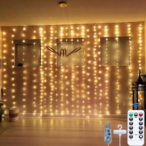 May include: A curtain of warm white LED string lights hanging against a wall, featuring multiple vertical strands of glowing bulbs. The set includes a USB connector, a small white hook, and a remote control with various numbered settings.