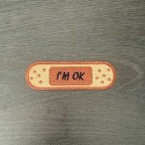 Puede incluir: Un parche bordado con forma de tirita. El parche es de color beige con un centro marrón más oscuro que dice "I'M OK" en letras marrones. El parche tiene pequeños puntos beige a cada lado.