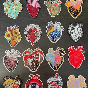 Op de afbeelding: Een verzameling geborduurde hartvormige patches in verschillende ontwerpen. De patches tonen anatomische harten, bloemstukken en unieke artistieke interpretaties. Kleuren zijn rood, roze, blauw, geel en zwart. Elke patch heeft een witte rand.