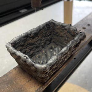 Könnte beinhalten: Ein rechteckiger Metallbehälter mit einer strukturierten, dunkelgrauen Oberfläche. Die Kanten sind leicht abgerundet und haben eine hellere graue Farbe. Der Behälter steht auf einer Holzoberfläche.