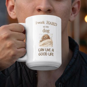 Può includere: Tazza in ceramica bianca con la scritta "I work HARD so my dog CAN LIVE A GOOD LIFE." La tazza presenta un'illustrazione ad acquerello di un cane che dorme sotto una coperta. La tazza è tenuta da una persona.