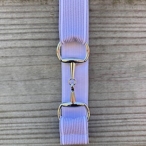 May include: A light purple and white striped horse bit cheek piece with silver-colored metal hardware. The cheek piece is made of a soft fabric and is designed to attach to a horse's bridle.