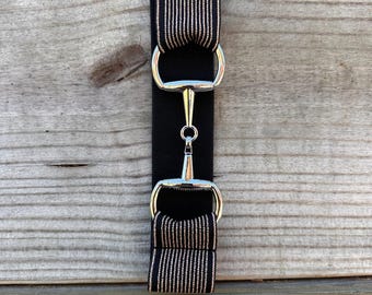 Pode incluir: Um cinto elástico preto com uma fivela de metal prateado em forma de bridão ao centro. A tira do cinto é composta por duas secções elásticas pretas com riscas verticais finas e claras, ligadas por um segmento elástico preto sólido.