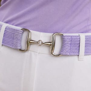 May include: A light purple belt with a silver bit buckle is worn over white pants. The belt has a striped pattern and white belt loops. The person is wearing a light purple shirt.
