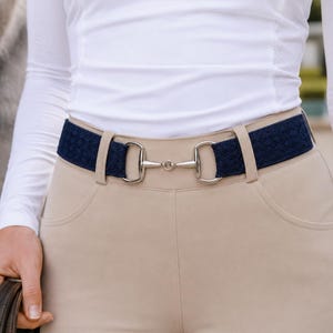 Könnte beinhalten: Ein marineblauer Reitgürtel mit einer silberfarbenen Gebiss-Schnalle. Der Gürtel wird mit beigefarbenen Reithosen und einem weißen Langarmshirt getragen. Der Gürtel hat ein gewebtes Muster.
