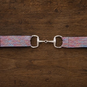 May include: A belt featuring a multicolored, textured fabric strap with a metallic silver-toned horse bit buckle. The strap displays a vibrant, speckled pattern of blue, pink, and purple hues, connected by a central silver-colored metal bit.