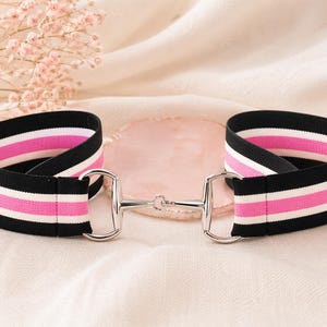 May include: An elastic belt featuring horizontal stripes in black, white, and bright pink. The belt is secured in the center with a silver-toned metal horse bit buckle, which connects the two ends of the striped fabric band together.