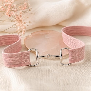 May include: A pink elastic belt with a silver-colored metal horse bit buckle. The belt is made of a textured fabric and is displayed on a cream-colored surface with a pink stone and pink flowers in the background.