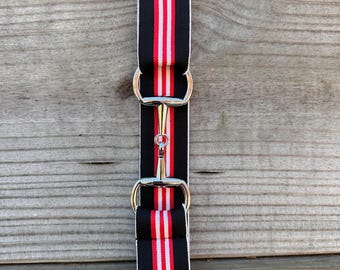 Pode incluir: Um cinto elástico com um padrão de riscas pretas, vermelhas e brancas. O cinto é fixado com uma fivela de metal prateado em forma de bridão, que liga duas secções da correia de tecido. O design é simétrico com um bridão central.