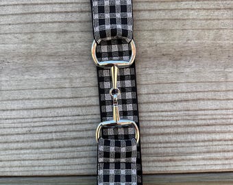 Pode incluir: Um freio de cavalo xadrez preto e branco com ferragens de metal prateado. O freio é feito de um tecido estampado e é projetado para uso equestre. O freio é mostrado em um fundo de madeira.