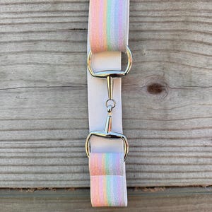 May include: An elastic belt featuring a pastel rainbow striped pattern on the ends and a solid white centre section. The belt is secured with a silver-toned metal bit-style buckle that connects the two fabric segments with a central horizontal bar.