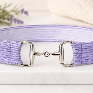 May include: A lavender elastic belt featuring a silver-toned metal horse bit buckle. The belt is constructed from a ribbed fabric material and includes two circular metal rings that connect the elastic band to the central bit hardware piece.
