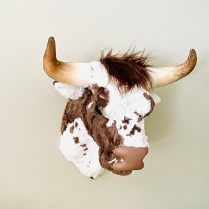 May include: A decorative bull head wall mount. The faux taxidermy piece features a white and brown spotted pattern, brown faux fur, and large, curved horns. The bull has a brown leather nose and is designed to be mounted on a wall.