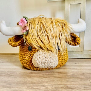 May include: A crocheted Highland cow head in shades of mustard yellow and beige. The cow has fluffy, light brown hair, white horns, and a small floral crown. The eyes are black buttons.