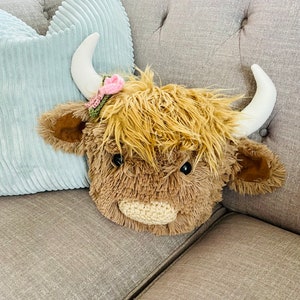 May include: A plush brown Highland Cow toy with a pink flower on its head. The toy is sitting on a gray couch.
